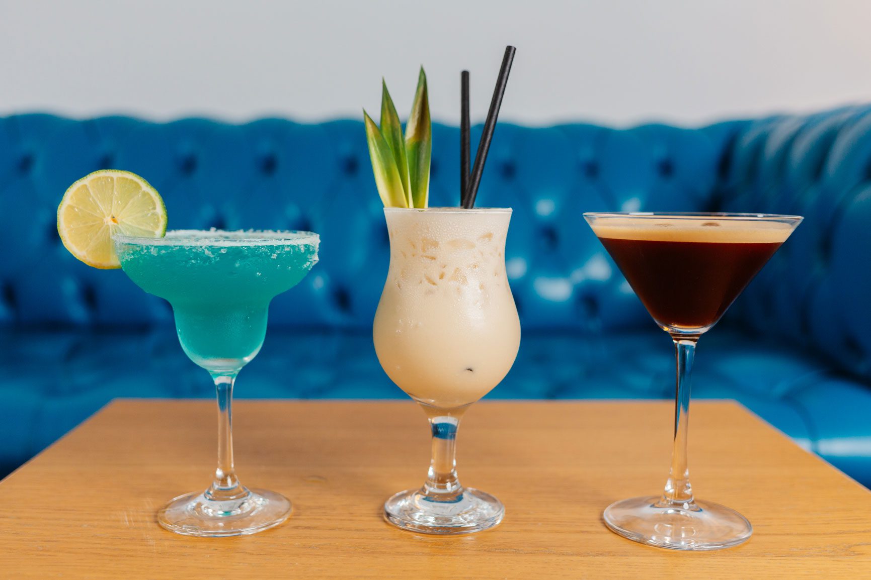 A blue lagoon, a pina colada and an espresso martini cocktail in a row on a table with a blue sofa behind